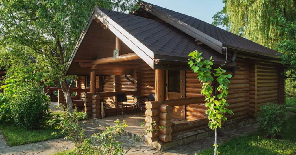A log cabin with a sidewalk leading up to it in a well-kept yard surrounded by trees and shrubs beneath a blue sky.