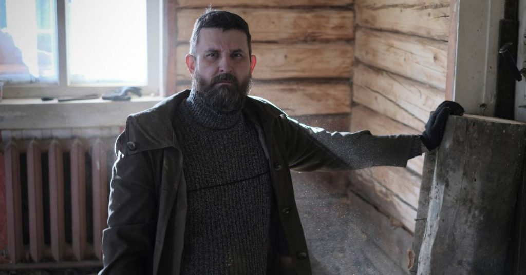 A man with a beard wearing a sweater, coat, and gloves standing in a cabin next to a large piece of wood.