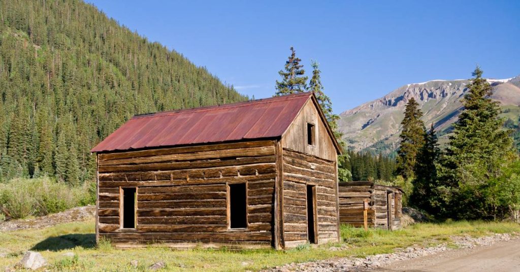 A log building with a red metal roof on the side of a dirt road in the country with mountains in the background.
