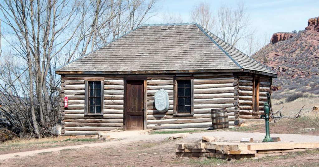 A small log home with a fire extinguisher and metal bucket hanging on the outside wall in front of a platform with a water pump.