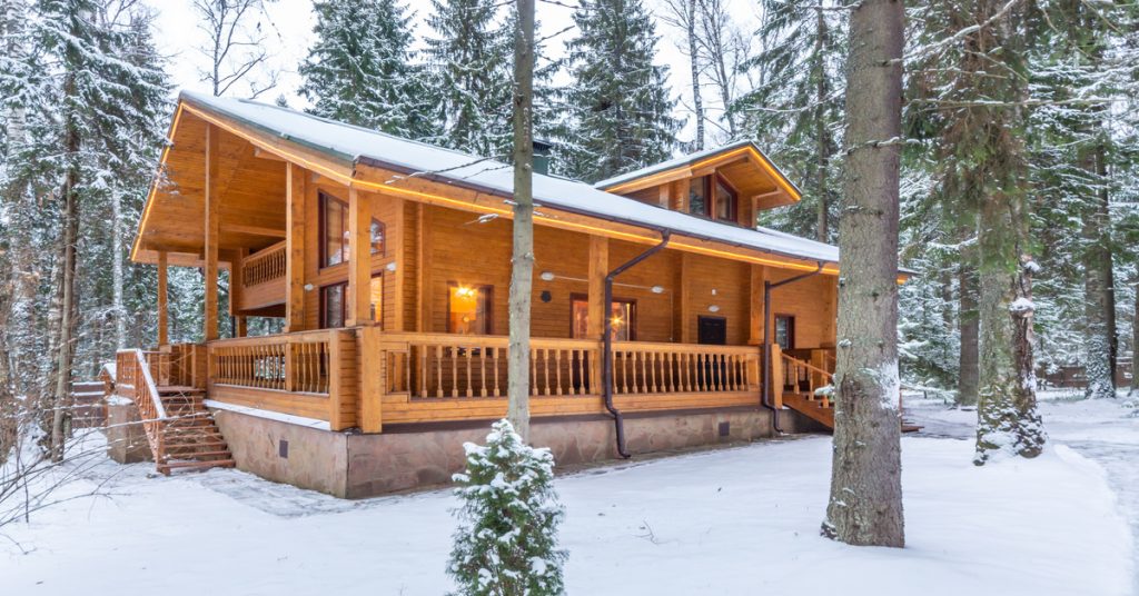 An outside view of a log cabin in the middle of the woods during the wintertime with freshly fallen snow around it.
